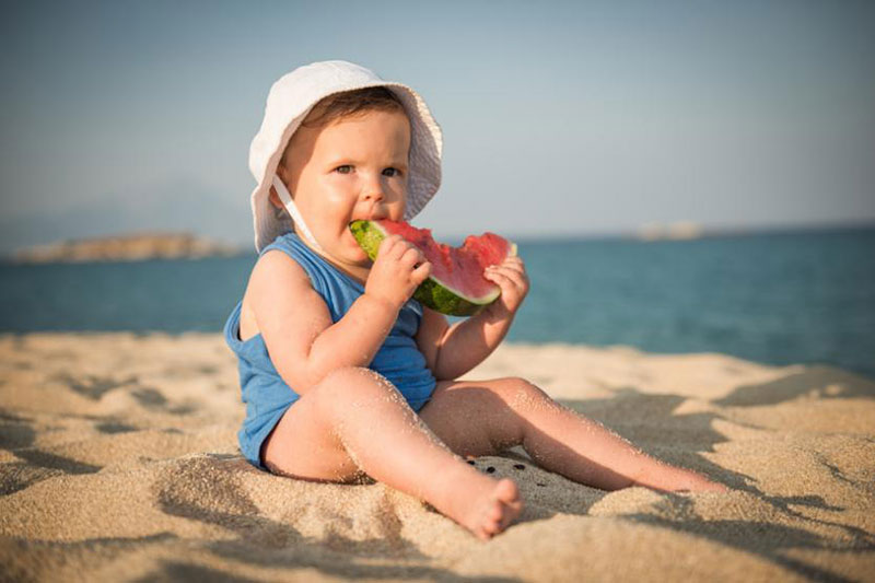 nina pequena con sombrero blanco y camiseta manga corta azul comiendo sandia en la playa