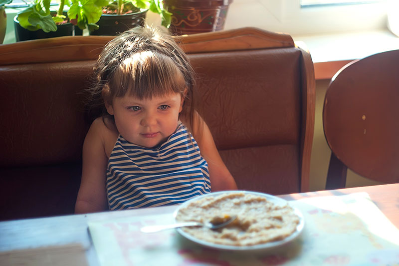 nina pequena que no quiere comer sentada en butaca mientras mira el plato de comida que tiene en la mesa