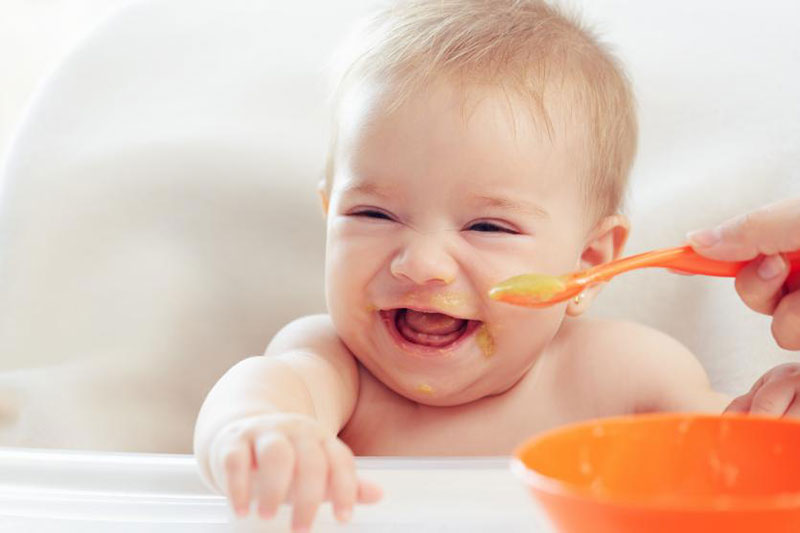 bebe comiendo en silla de comer sin ropa muy sonriente mientras le dan papilla con bol y cuchara naranja