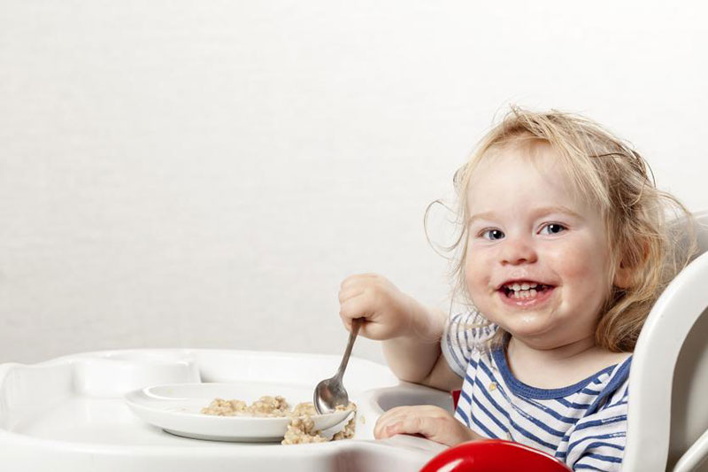 nina pequena en silla de comer comiendo sola con plato y cuchara sonriendo a cámara